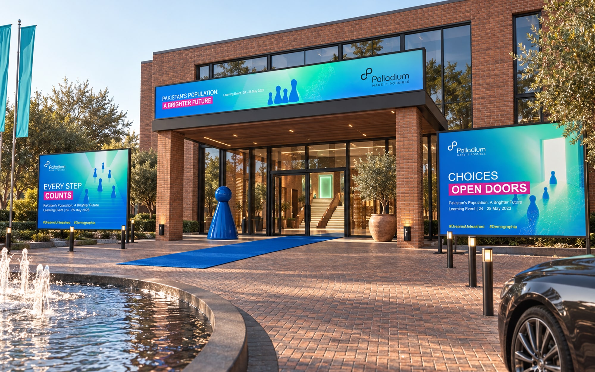 The entrance of a brick building with a glazed front: a long banner with the event title along the top of the canopy, a blue carpet to the doors, one blue pawn sculpture beside them, a billboard on a frame either side