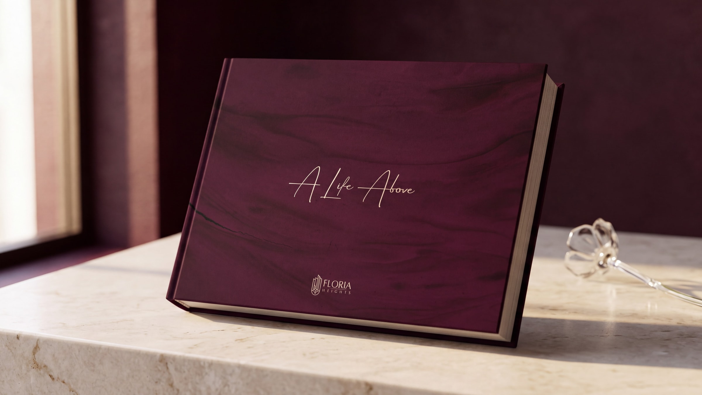 The Floria Heights sales book closed on cream marble, its wine cover carrying a single line of champagne script and a small line-drawn mark, a glass flower stem beside it