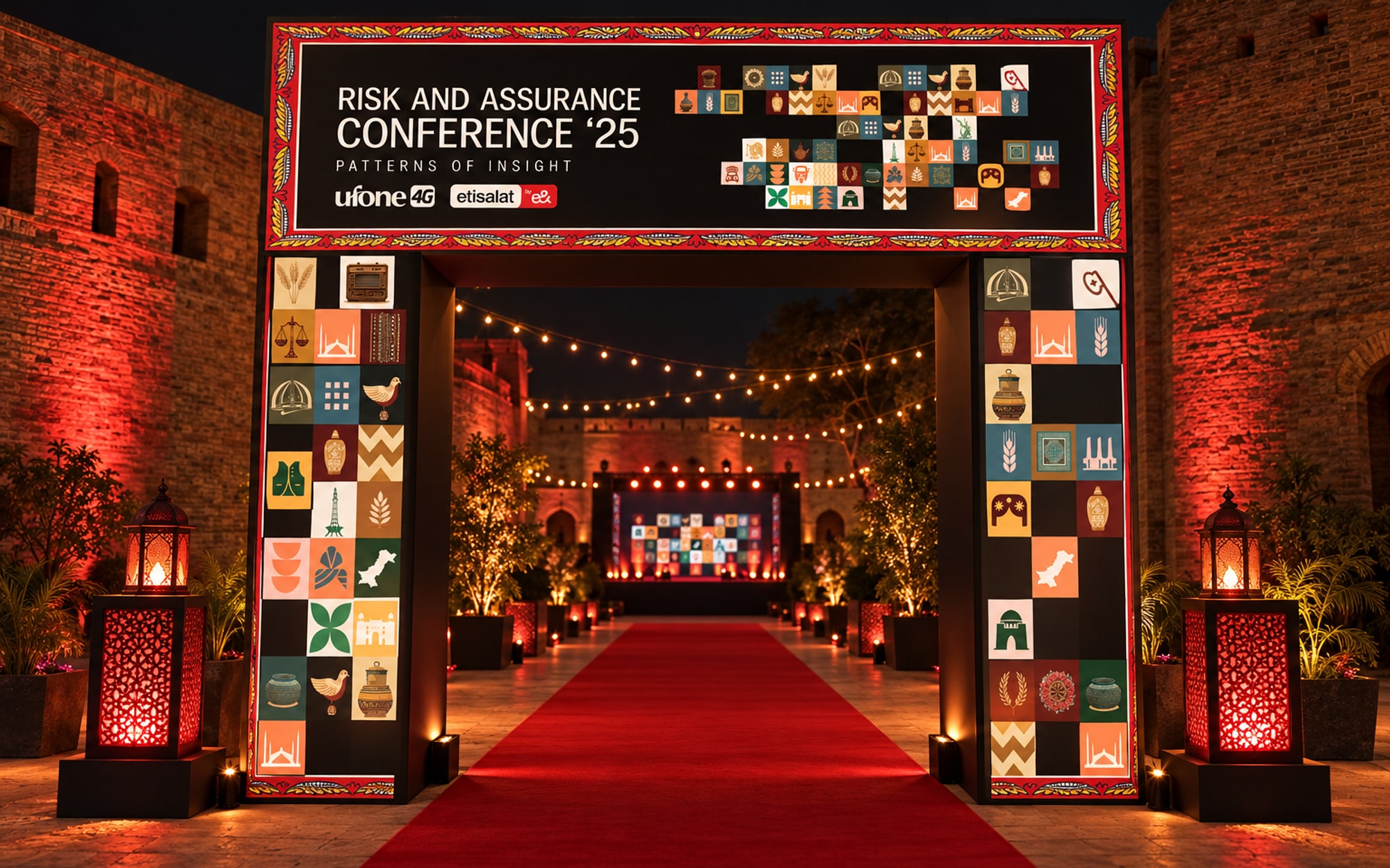 A black entrance portal at a brick fort at night, its header carrying the title and its legs covered in motif tiles, a red carpet running through it towards a lit stage