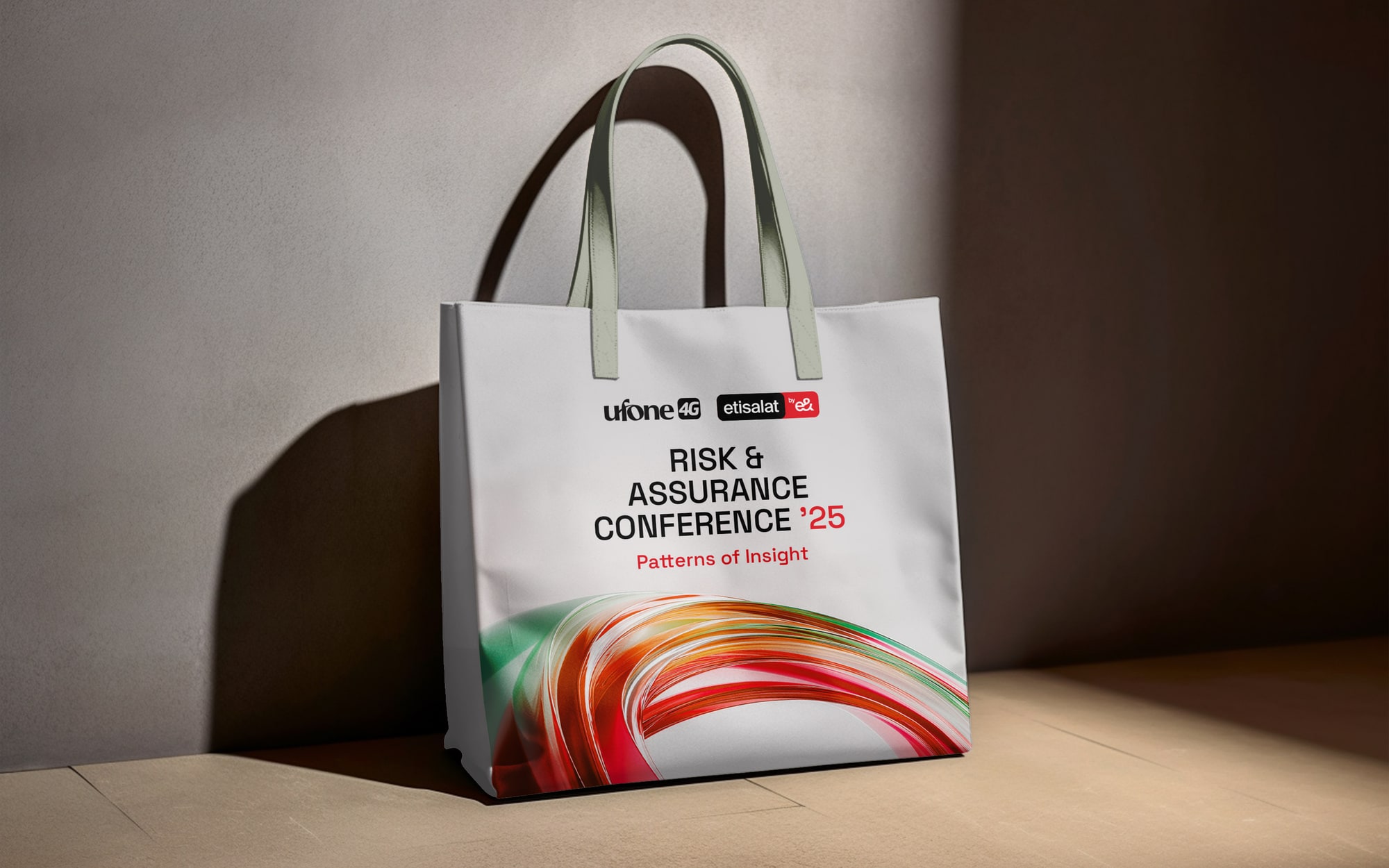 A white canvas tote printed with the conference title and the ribbon, standing against a plaster wall in raking light