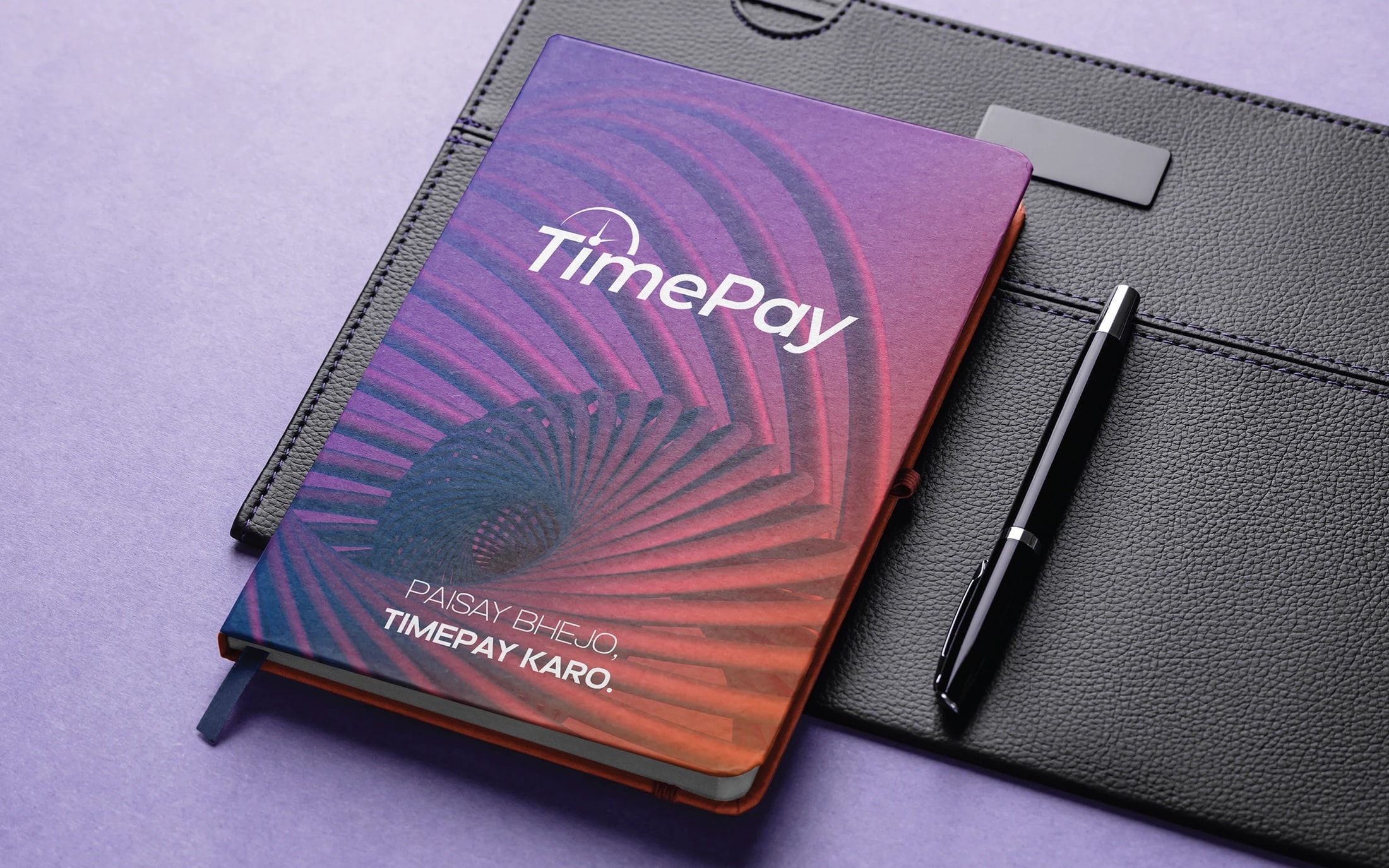 A hardback notebook with a gradient cover, the white logotype and the tagline, on a black leather folio with a pen