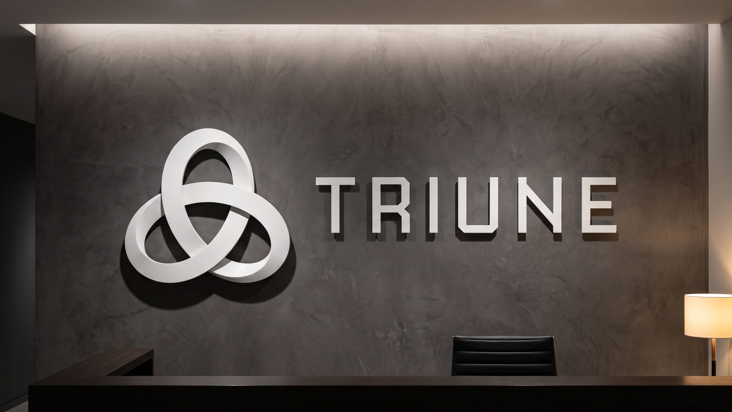 A reception desk under a dark plaster wall carrying the dimensional Triune sign, the woven symbol and the flat-cut wordmark both standing proud and lit from above